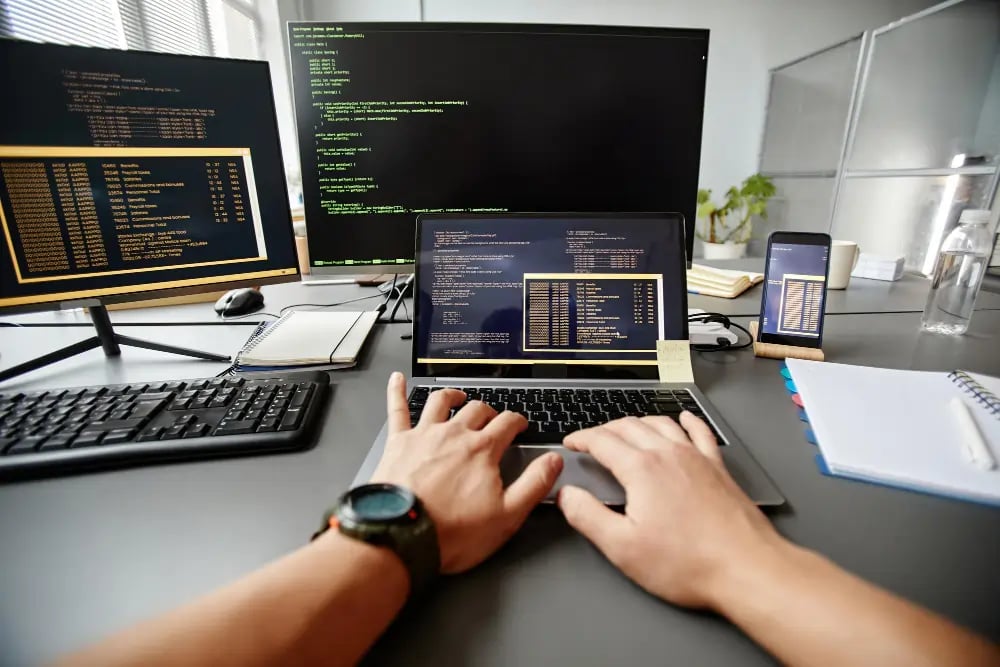 Man typing on laptop and two monitors showing some code