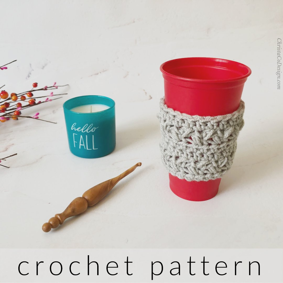 Red travel coffee cup with grey crochet sleeve next to hook, and candle.