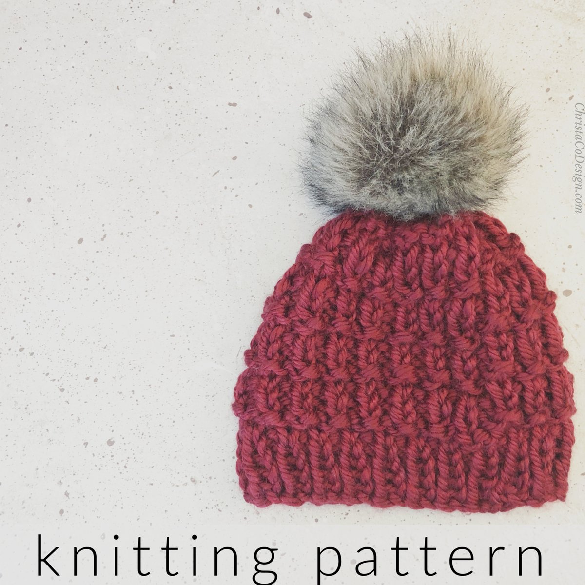 Burgundy textured knit hat with grey faux fur pom on grey background.