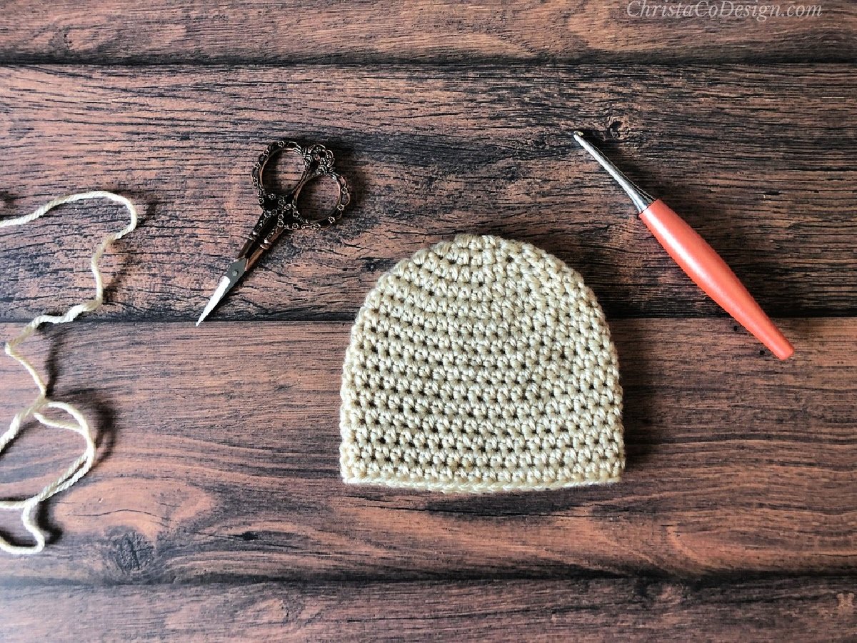 Crochet baby beanie in cream yarn with hook and scissors.