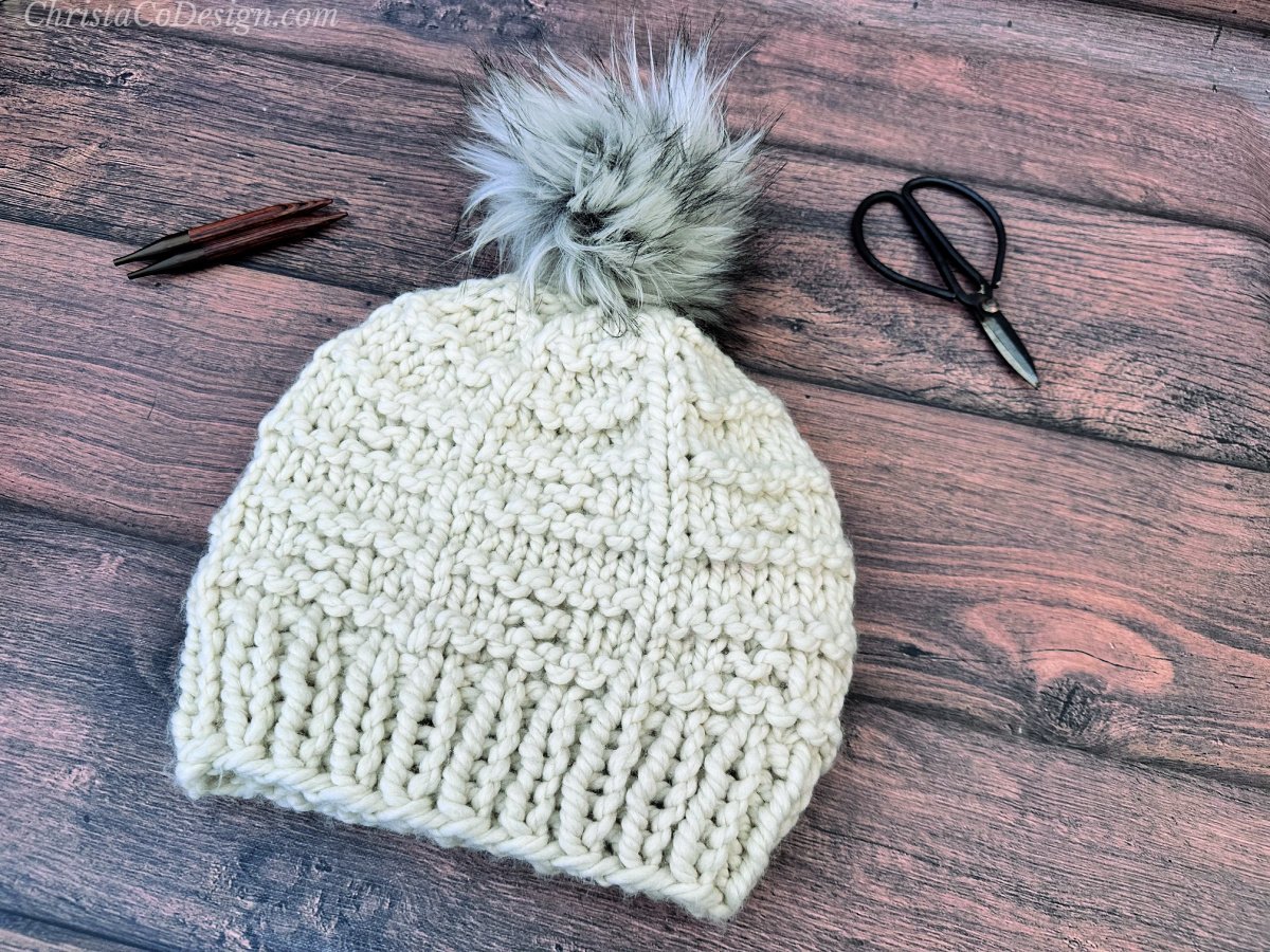 Textured knit hat in off white with grey pom laid flat with scissors and needles.