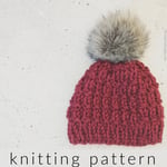 Burgundy textured knit hat with grey faux fur pom on grey background.