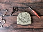 Crochet baby beanie in cream yarn with hook and scissors.