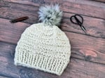 Textured knit hat in off white with grey pom laid flat with scissors and needles.