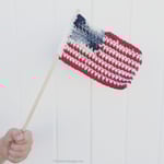 Hand holding crochet flag on dowel in front of white fence.