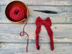 Red crochet bow with long tail ribbon next to yarn and hook.