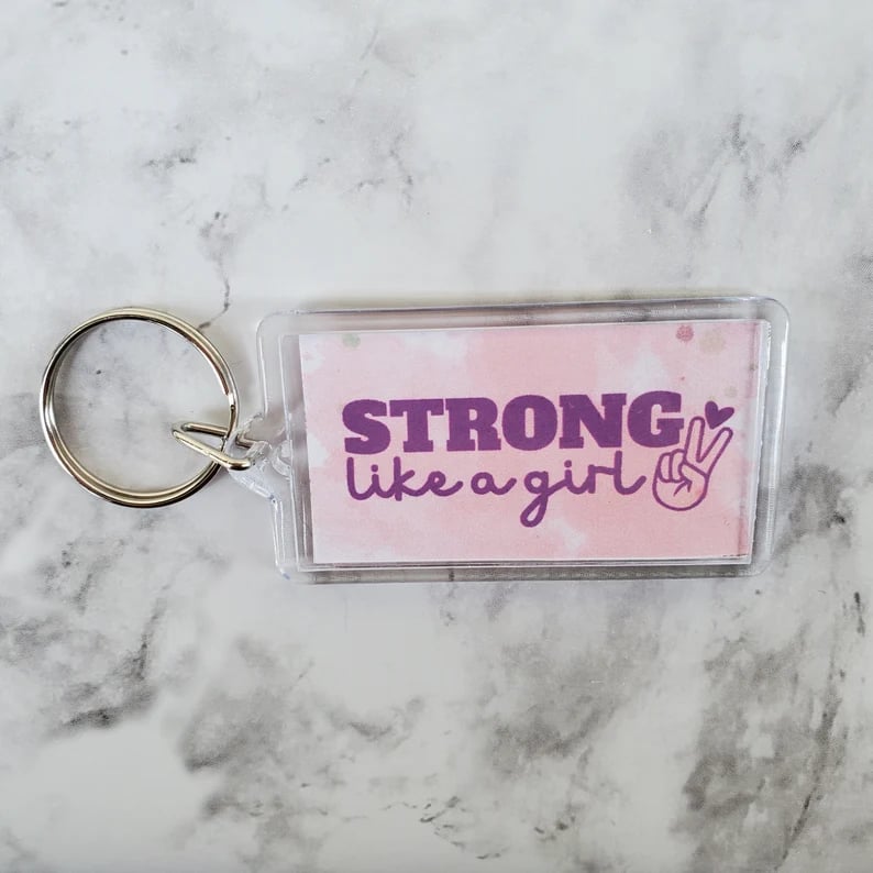 acrylic keychain pink with purple text saying strong like a girl, on a marble background