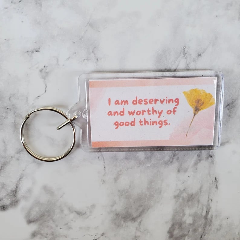 Keychain sitting on a marble surface. It is pink and white with a dried flower and pink lettering saying I am deserving and worthy of good things