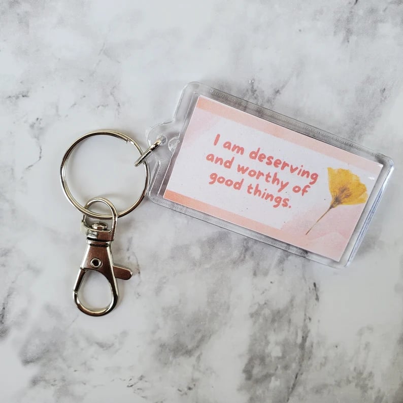 Keychain sitting on a marble surface. It is pink and white with a dried flower and pink lettering saying I am deserving and worthy of good things