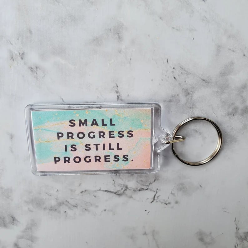 Keychain wiht teal and pink background that says small progress is still progress, sitting on a marble surface