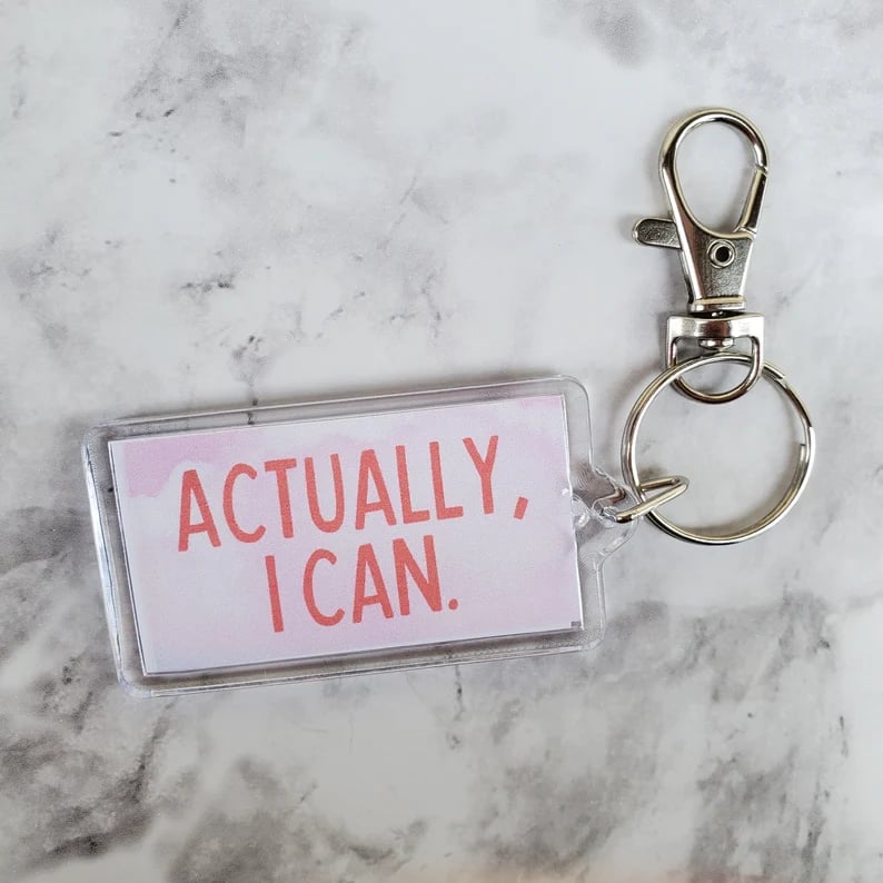 keychain with pink lettering saying Actually I can on a light pink background, sitting on a marble surface