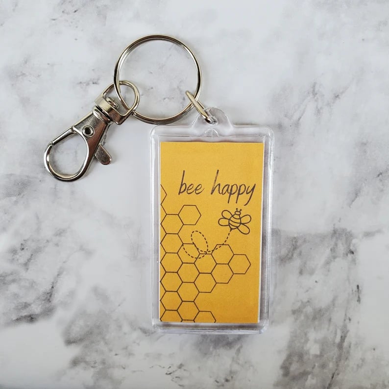 Keychain with yellow background, bee happy, a bee and a honeycomb, all on a marble surface