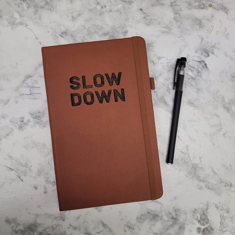 PU Brown Leather Journal with the words slow down burned into the cover sitting on a marble table with a black pen